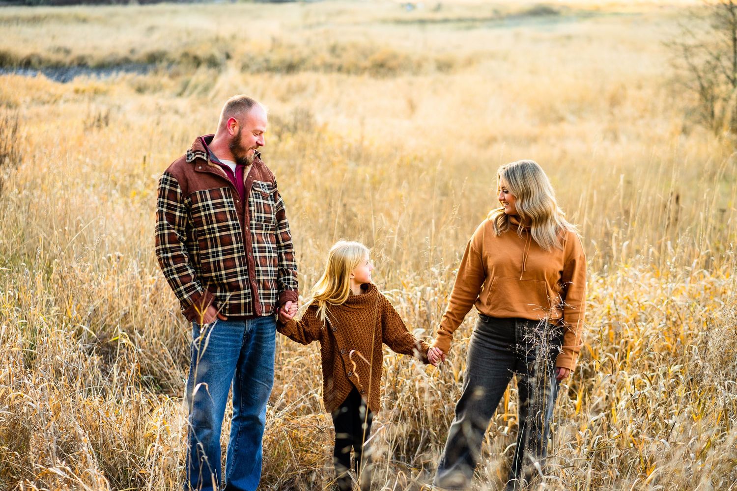 Happy family of three holding hands walking through golden autumn meadow at sunset.