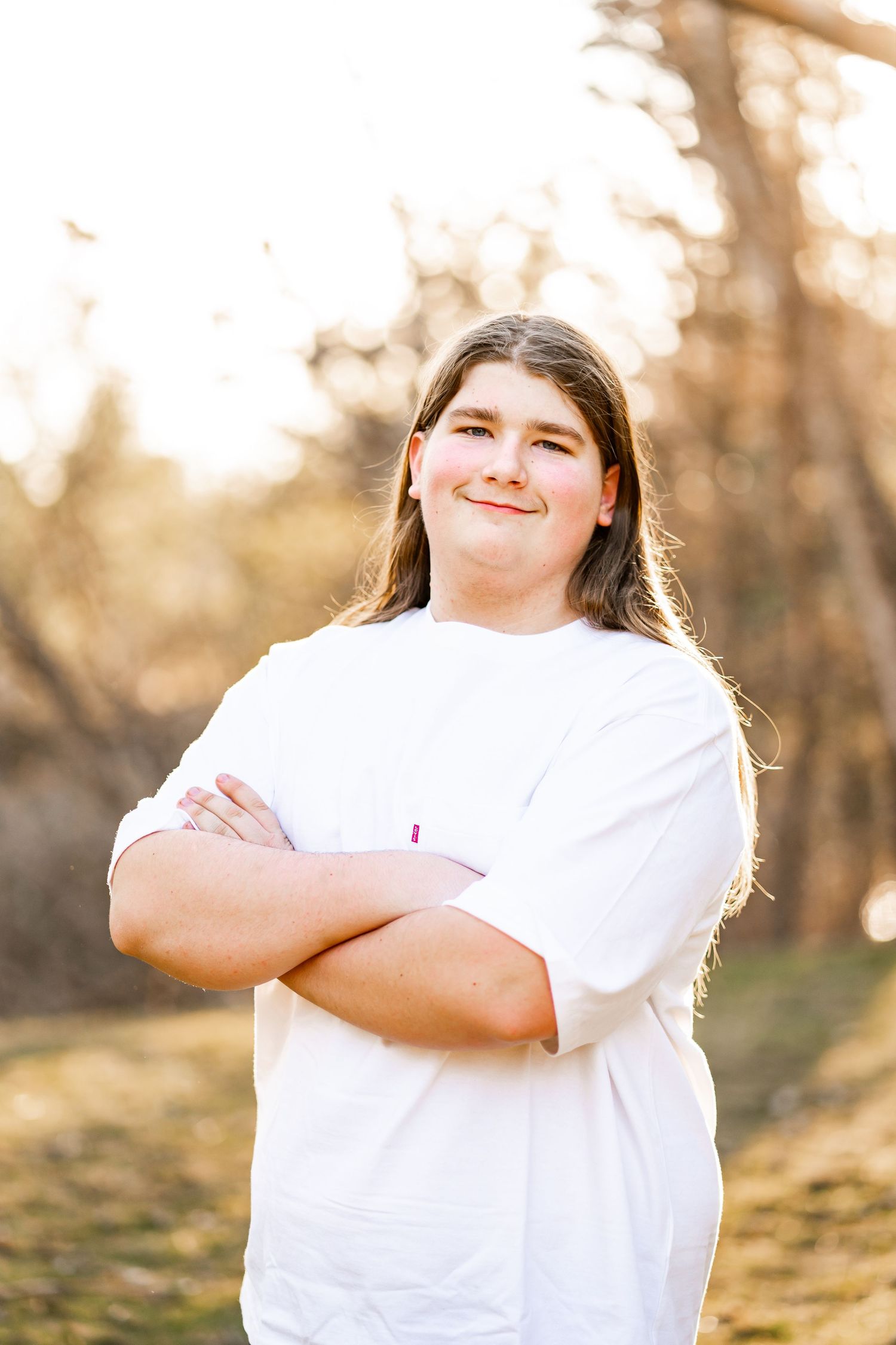 Teen with long hair standing arms crossed smiling in sunny outdoor setting.