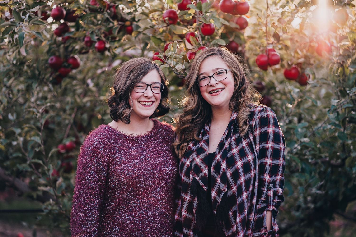Two women wearing glasses smiling together in front of red apple trees at sunset.