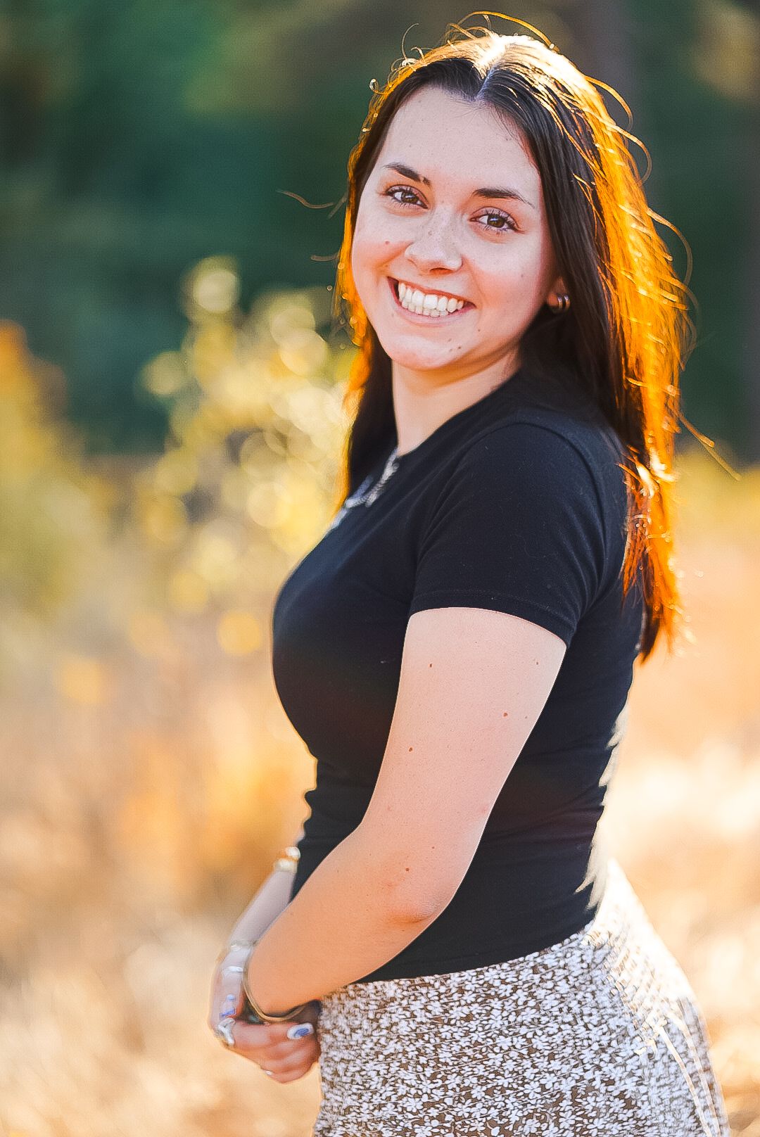 Young woman with dark hair smiling in black top with autumn bokeh background.