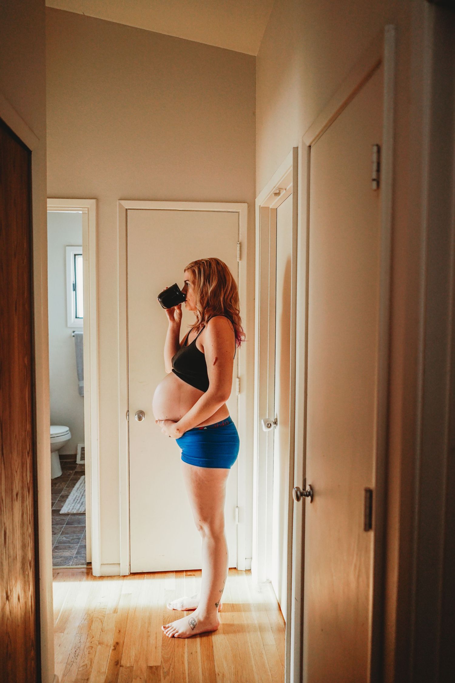 Pregnant woman in black bralette and blue shorts drinking from a mug in a hallway.