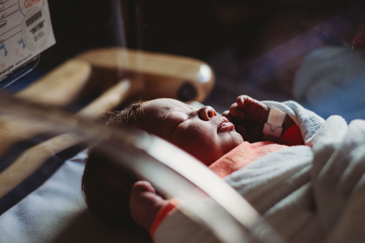 Newborn baby resting peacefully in a hospital bassinet under soft dramatic lighting.