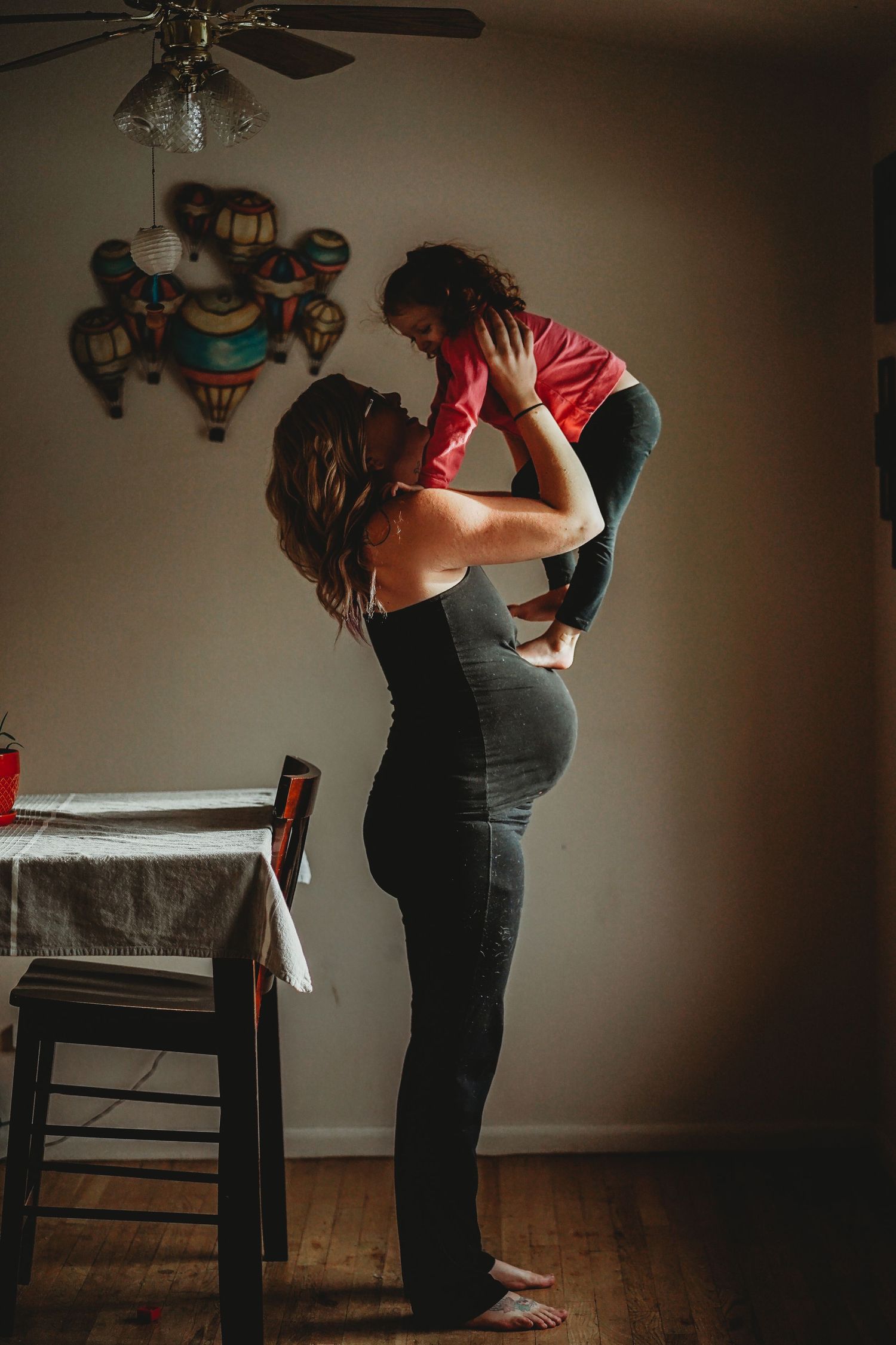 Mother lifting smiling toddler in red outfit above her head in a warmly lit living room.