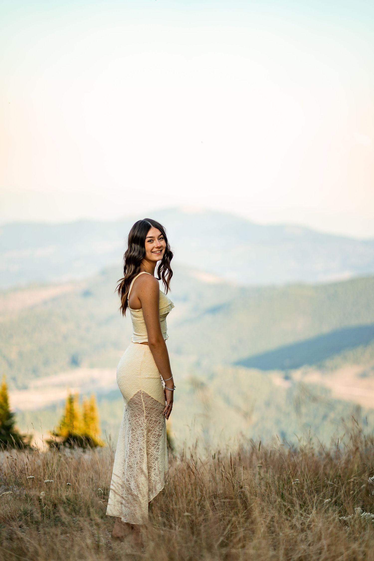 Young woman in cream lace skirt smiling over shoulder with scenic mountain valley backdrop.