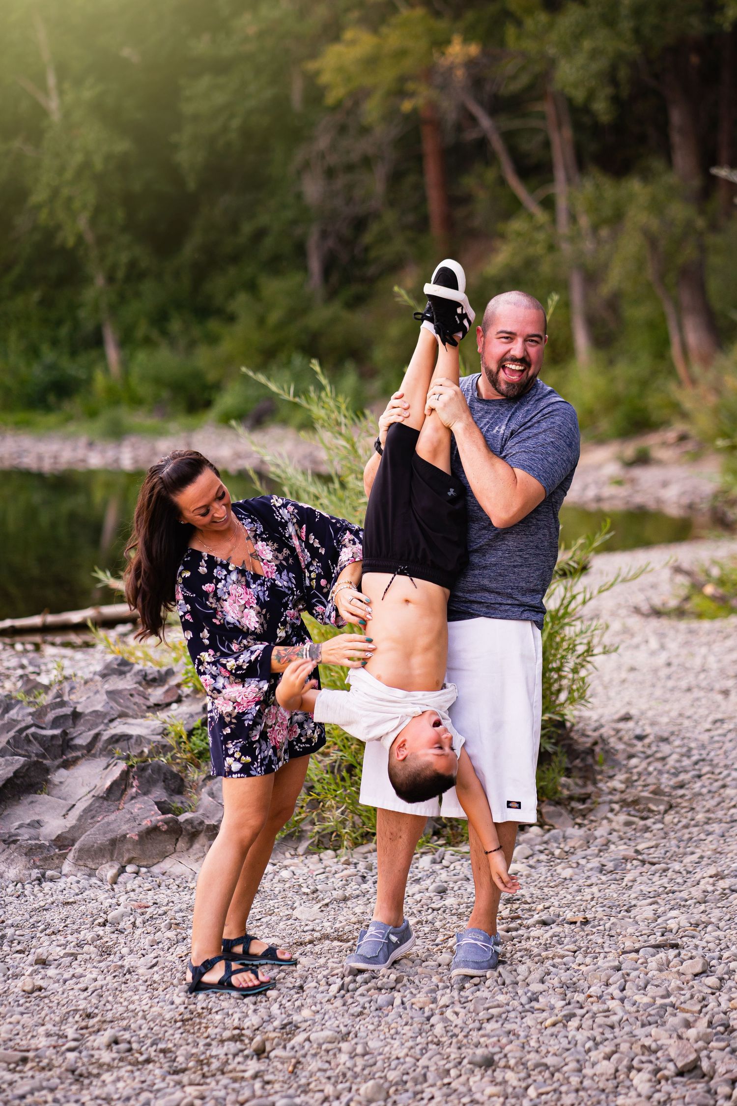 Parents laughing while playfully holding a child upside down on a rocky riverside.