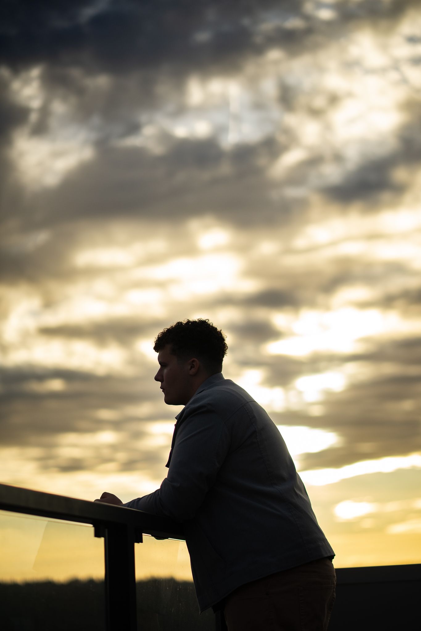 Silhouette of a young man leaning on a railing against a dramatic golden sunset sky with dark clouds.