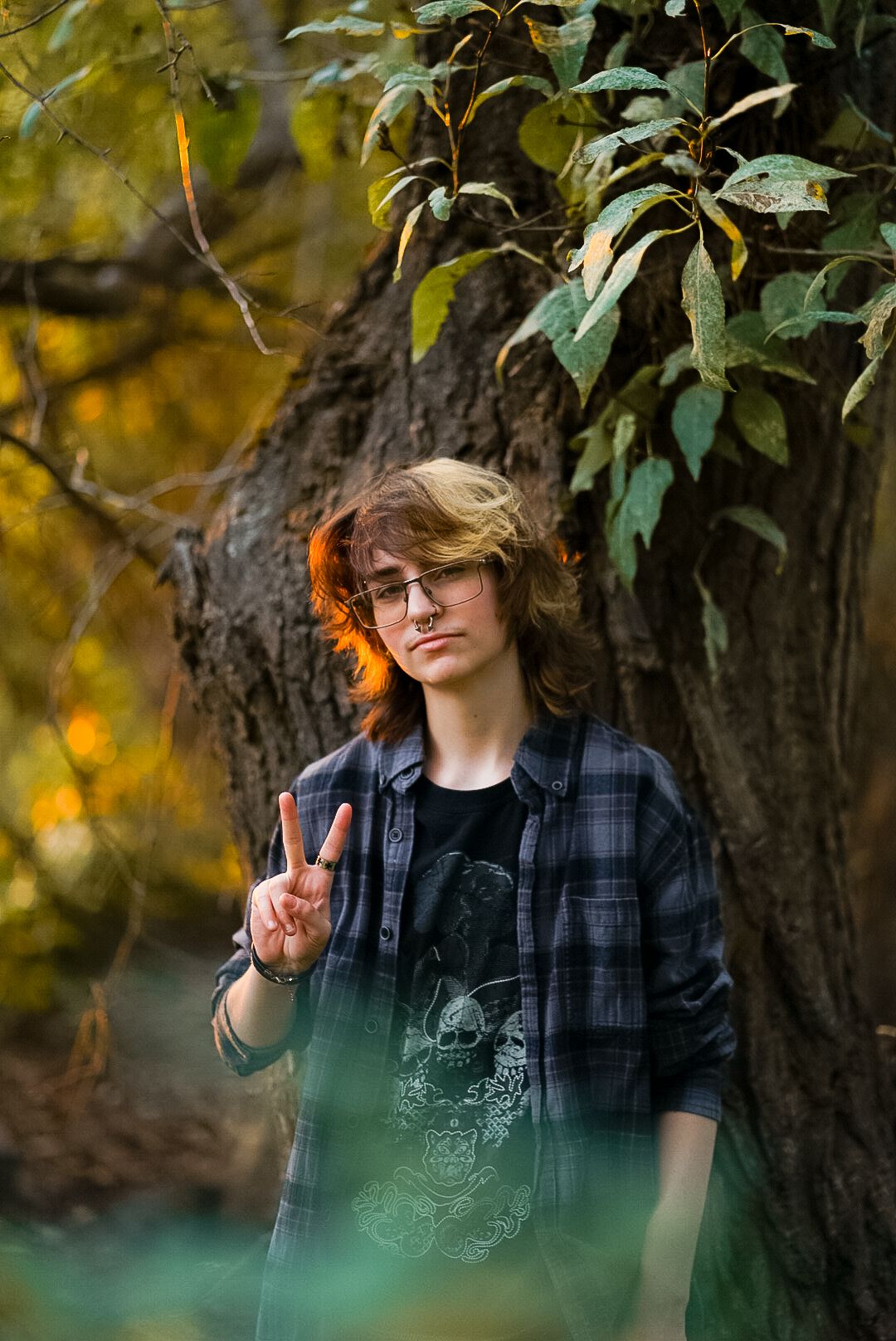 Teen with glasses giving peace sign leaning against tree in autumn forest wearing plaid shirt.