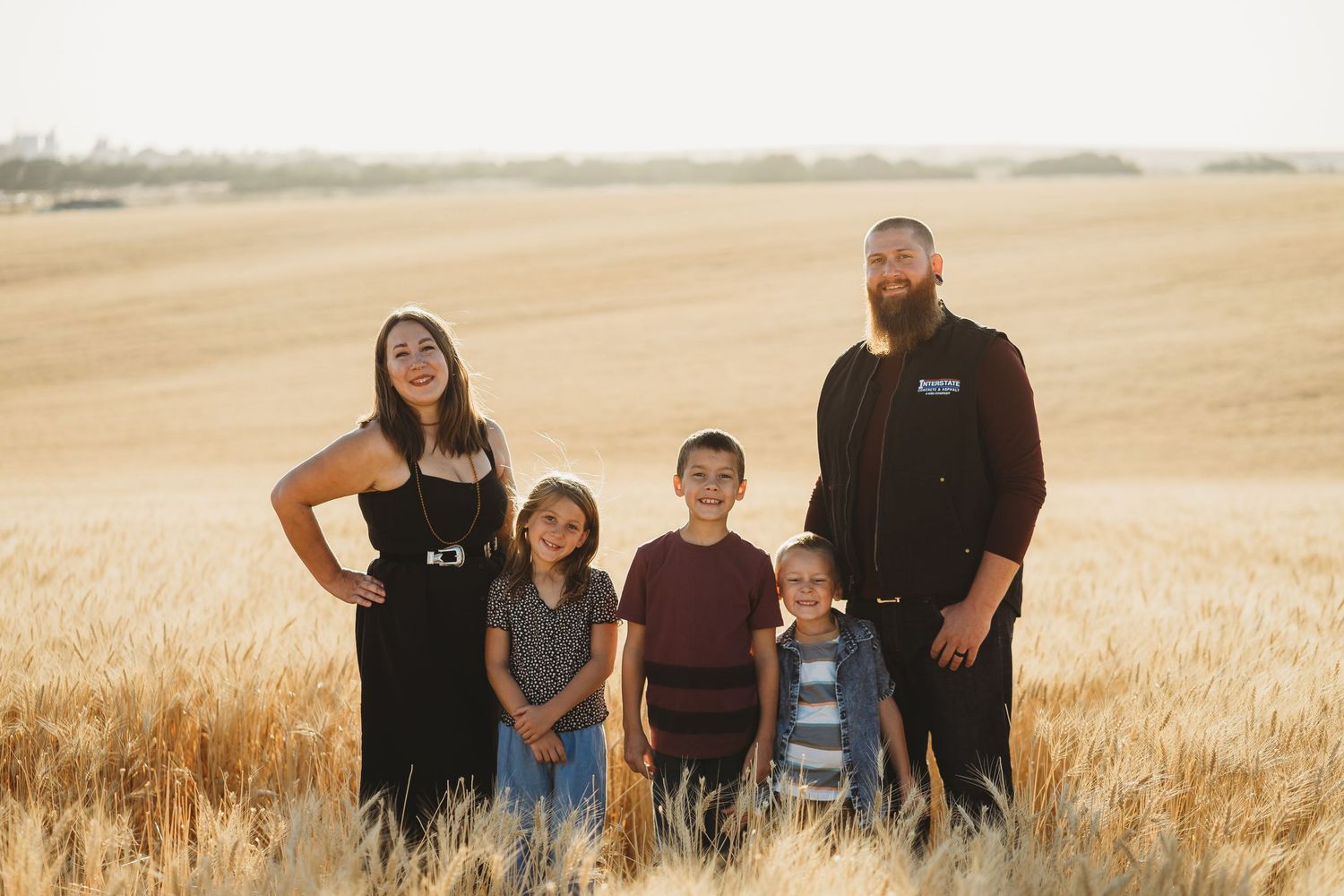 Family of five standing together in a golden wheat field at sunset.