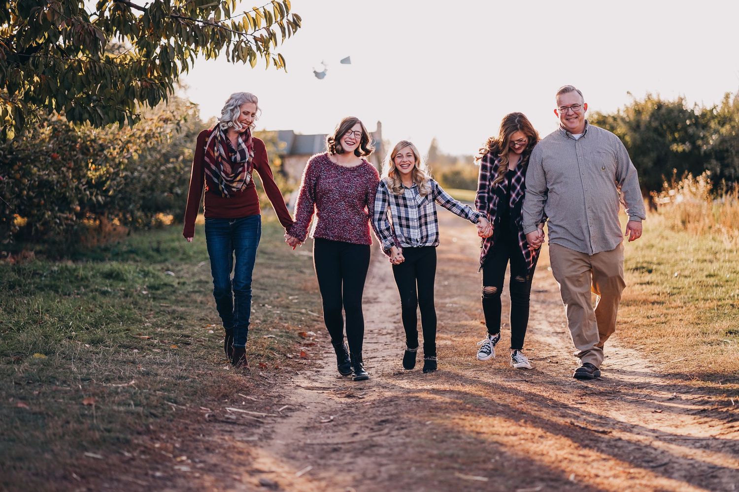 Family of five holding hands walking down a sunny dirt path through an autumn orchard.