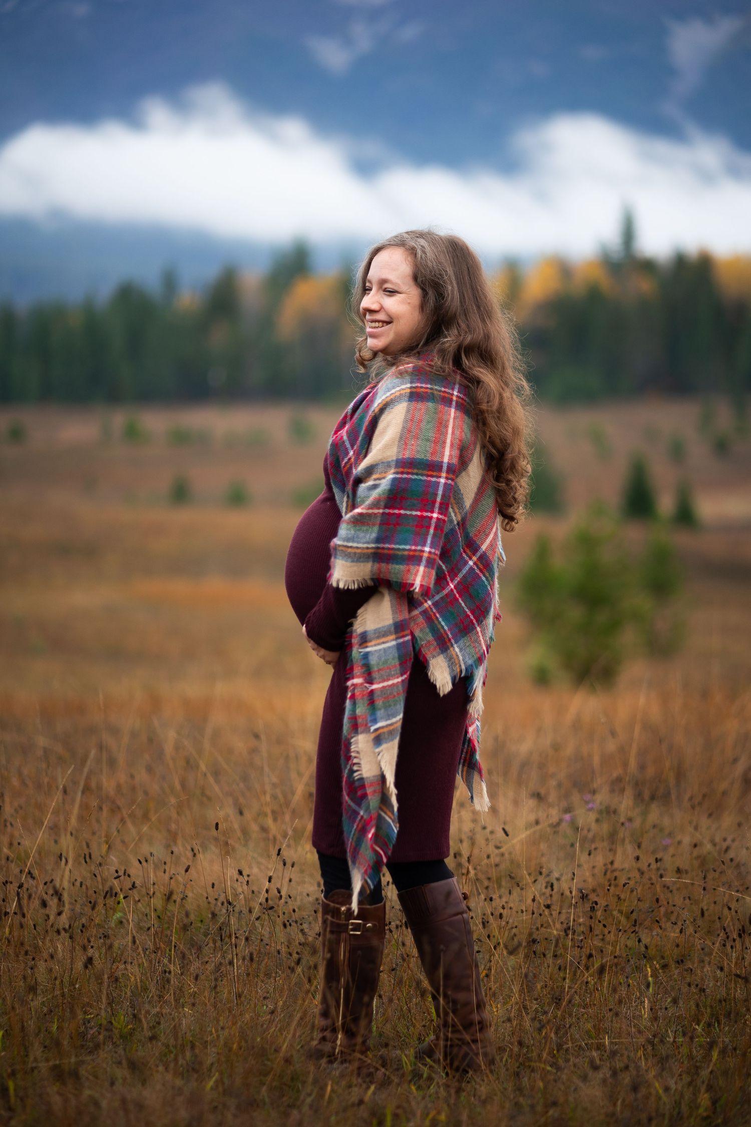 Smiling pregnant woman in plaid wrap standing in autumn field with mountain backdrop.