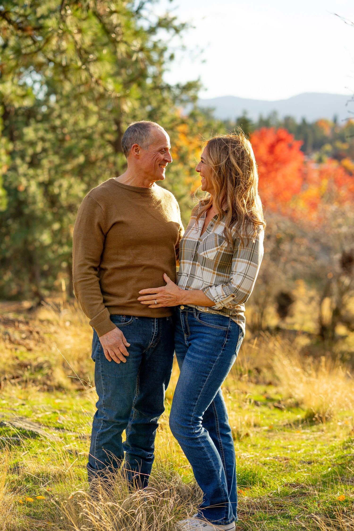 Couple smiling at each other in colorful autumn orchard with mountain backdrop.