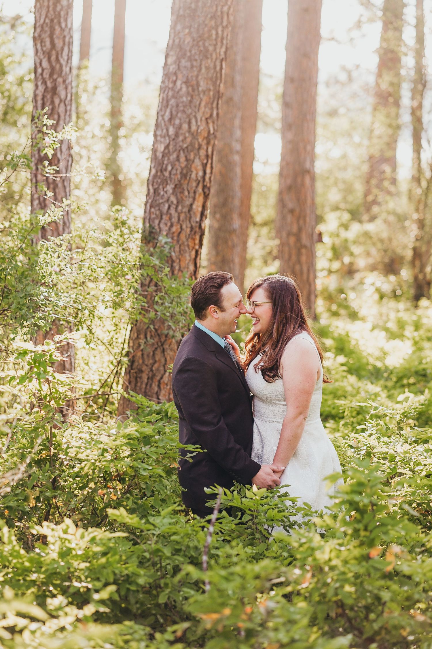 Couple sharing a romantic moment in a sunlit forest, dressed in wedding attire among lush green foliage.
