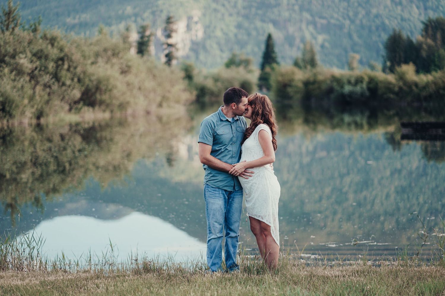 Couple sharing a kiss near a serene mountain lake with lush green forest backdrop.