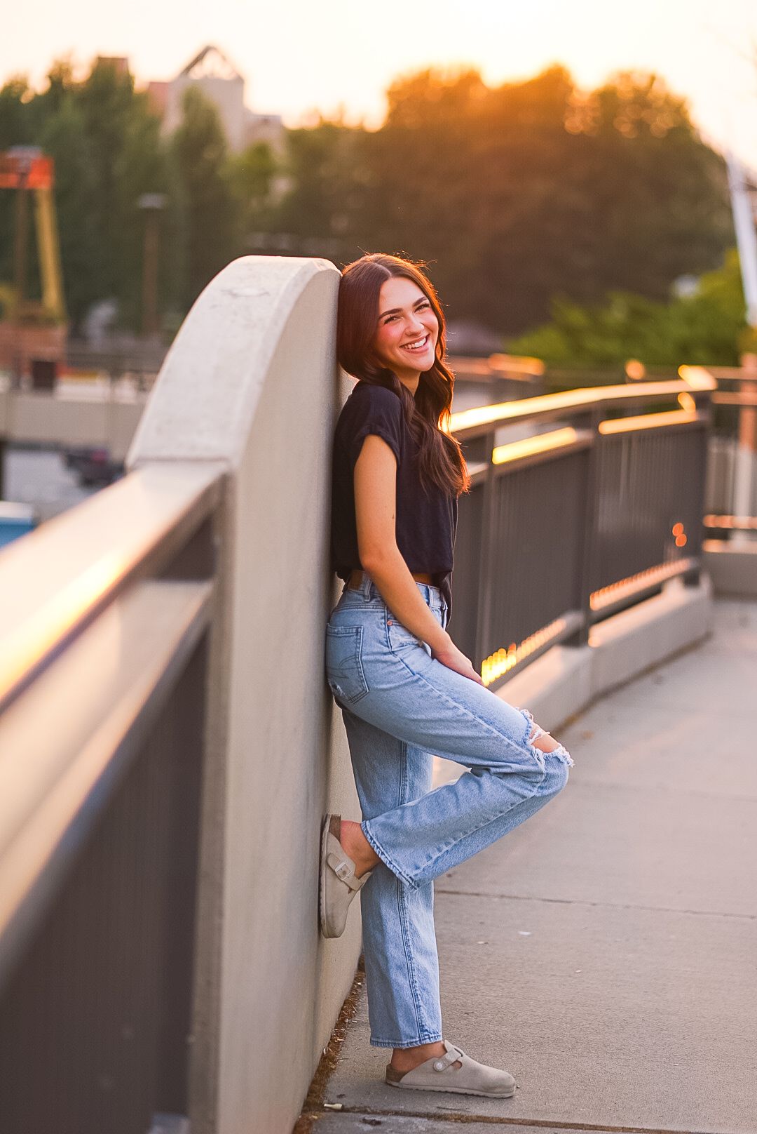 Laughing young woman in black top and jeans leaning against concrete railing at sunset.