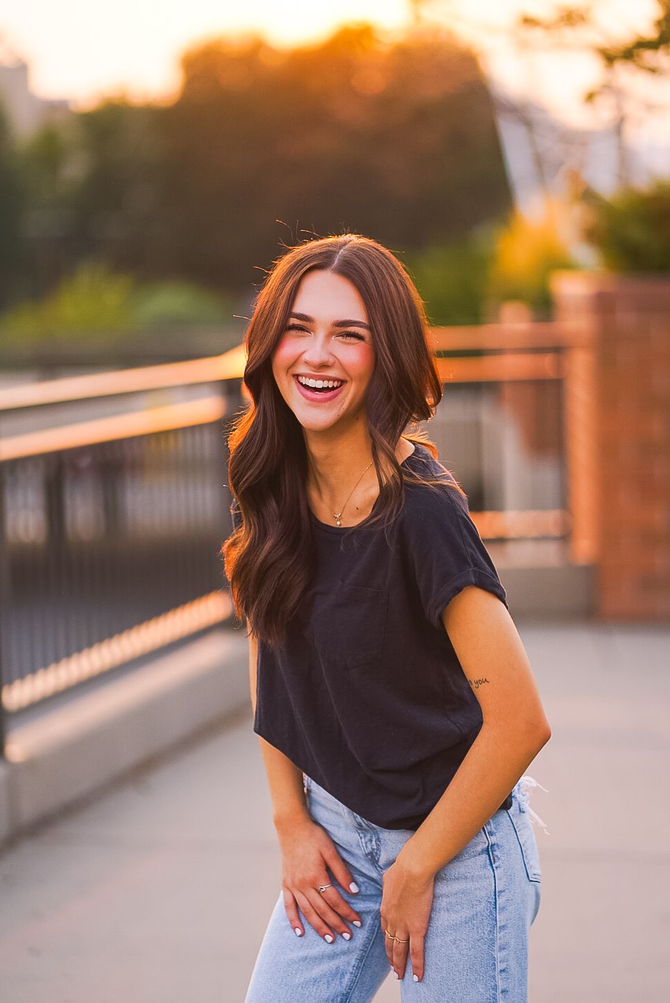 Laughing young woman in black t-shirt and jeans posing on bridge at warm golden sunset.