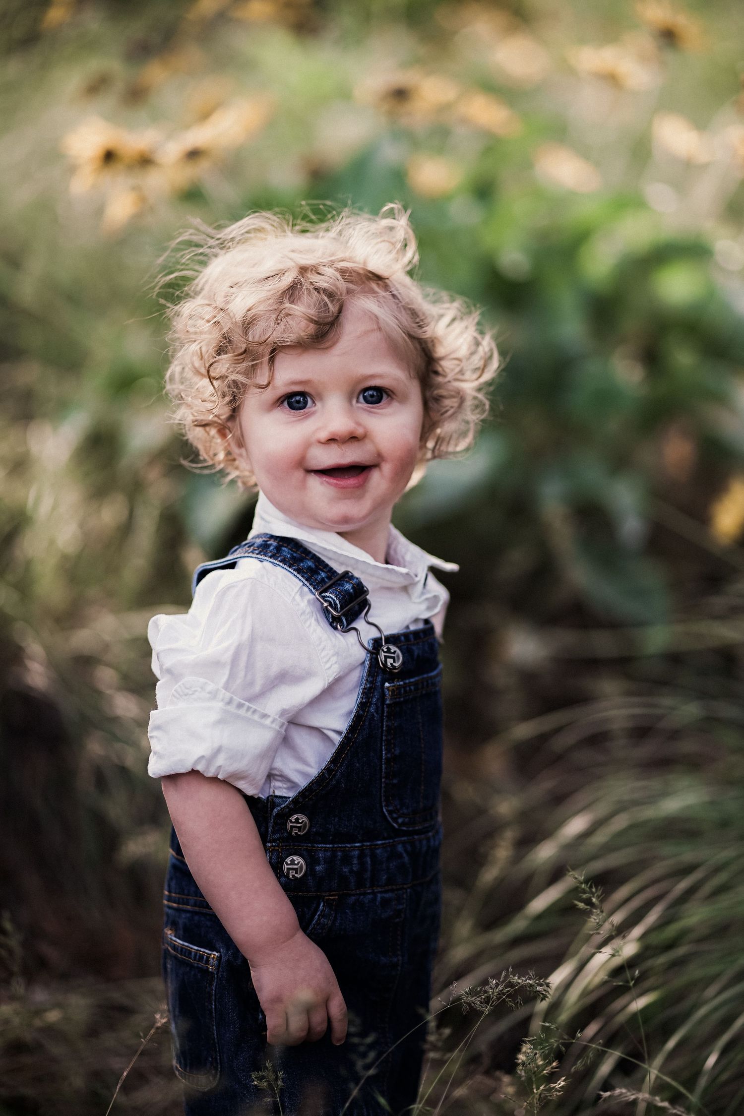 Adorable curly-haired toddler smiling in denim overalls and white shirt in a sunny garden.