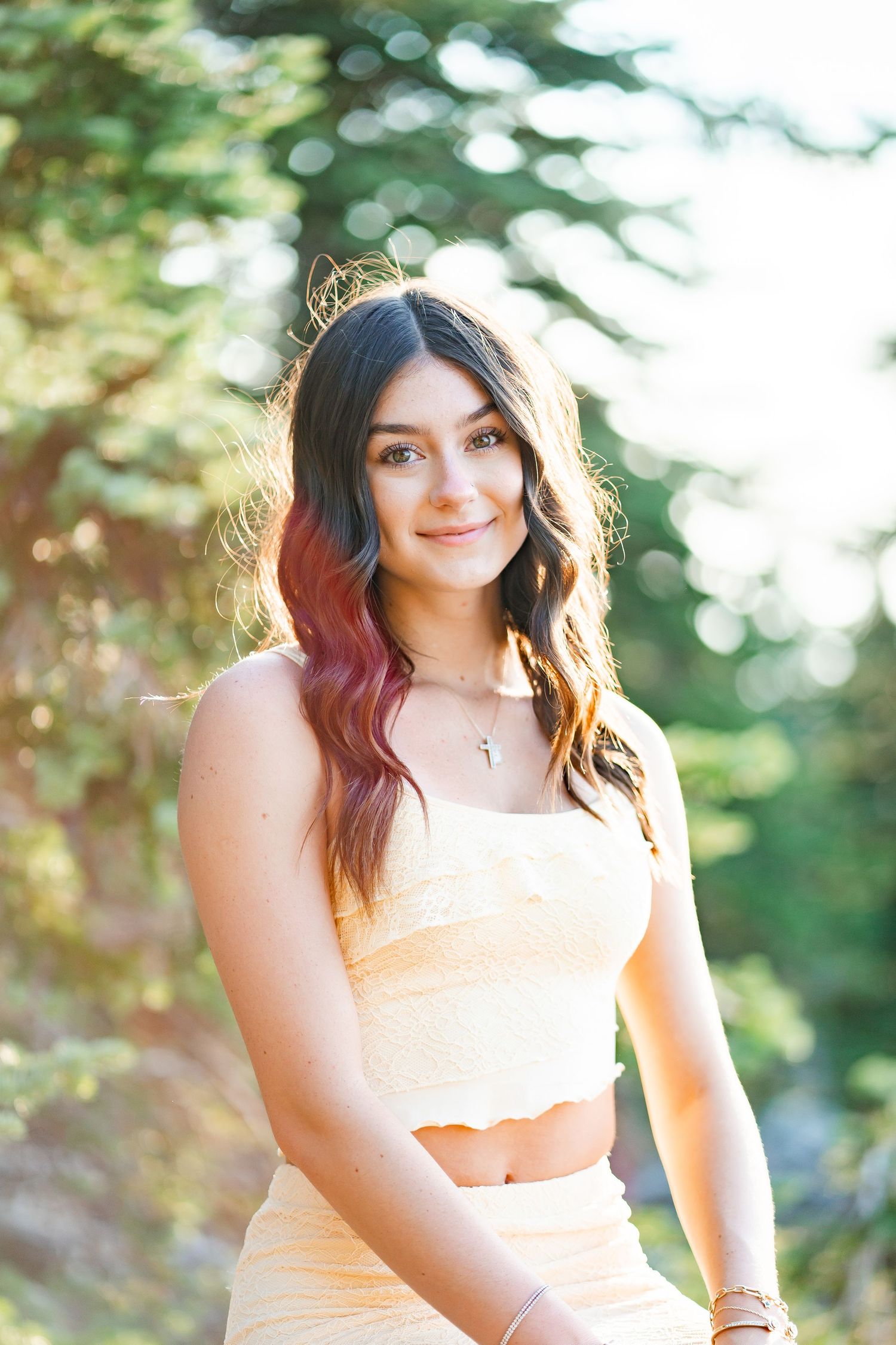 Smiling teen girl with pink highlighted hair in yellow outfit among pine trees.