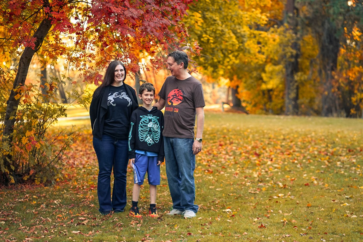 Family of three smiling together outdoors among vibrant red and orange autumn trees.