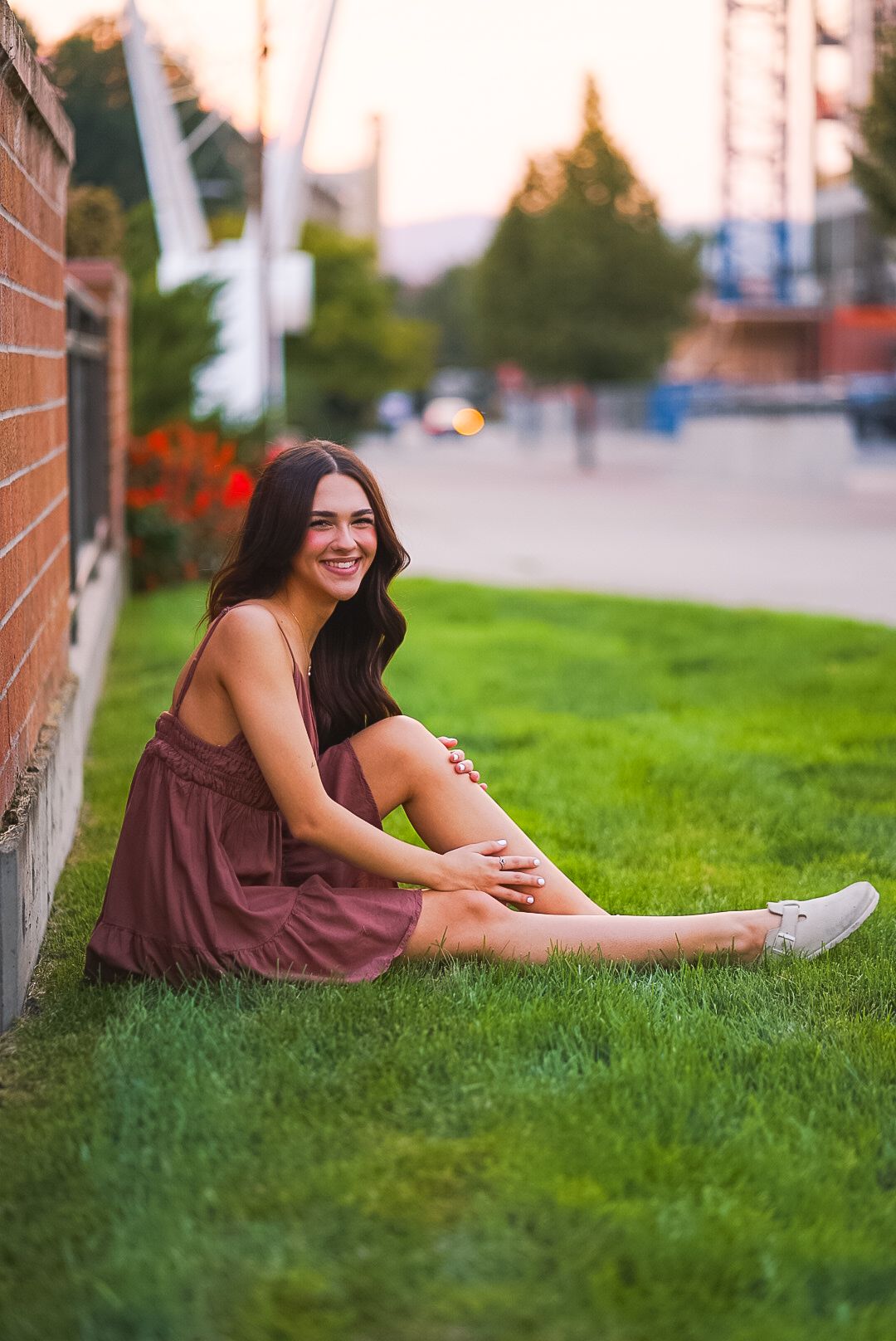 Smiling young woman in burgundy dress sitting on green grass at sunset.