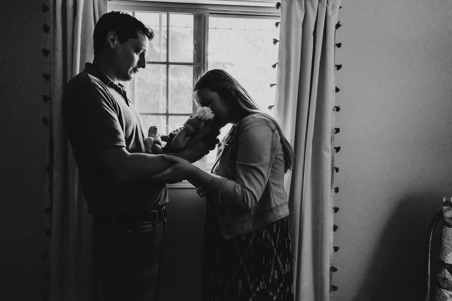 Black and white photo of parents tenderly holding newborn baby by window light.