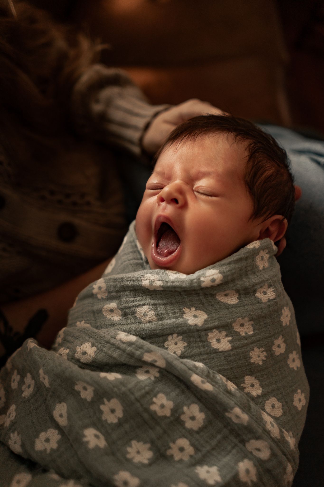 Newborn baby yawning while swaddled in a light blue floral muslin blanket, held by a person in warm indoor lighting.