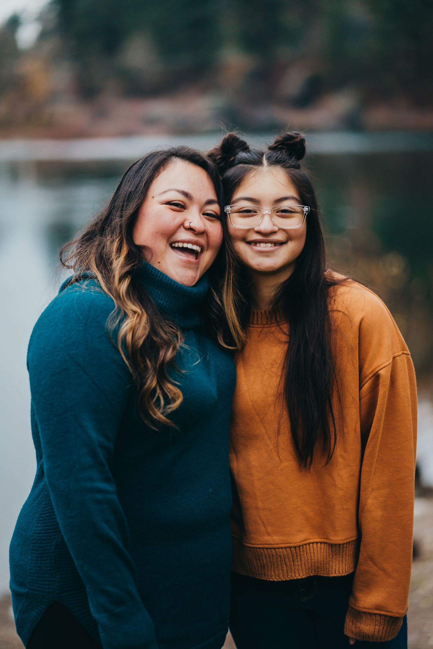 Two young women smiling and laughing together outdoors near a blurred lake background.