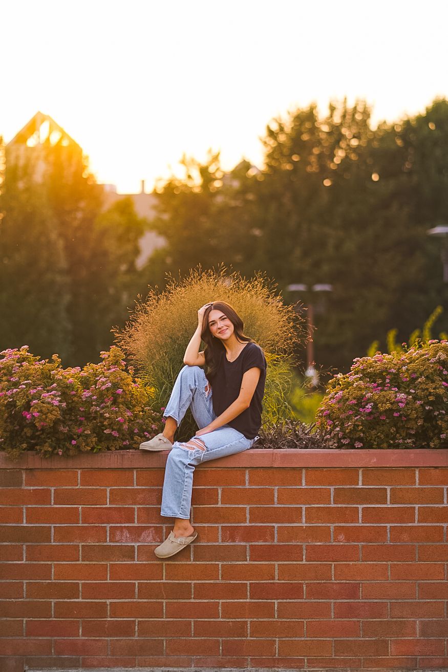 Teenage girl in black tee and jeans sitting on brick wall surrounded by flowers at sunset.