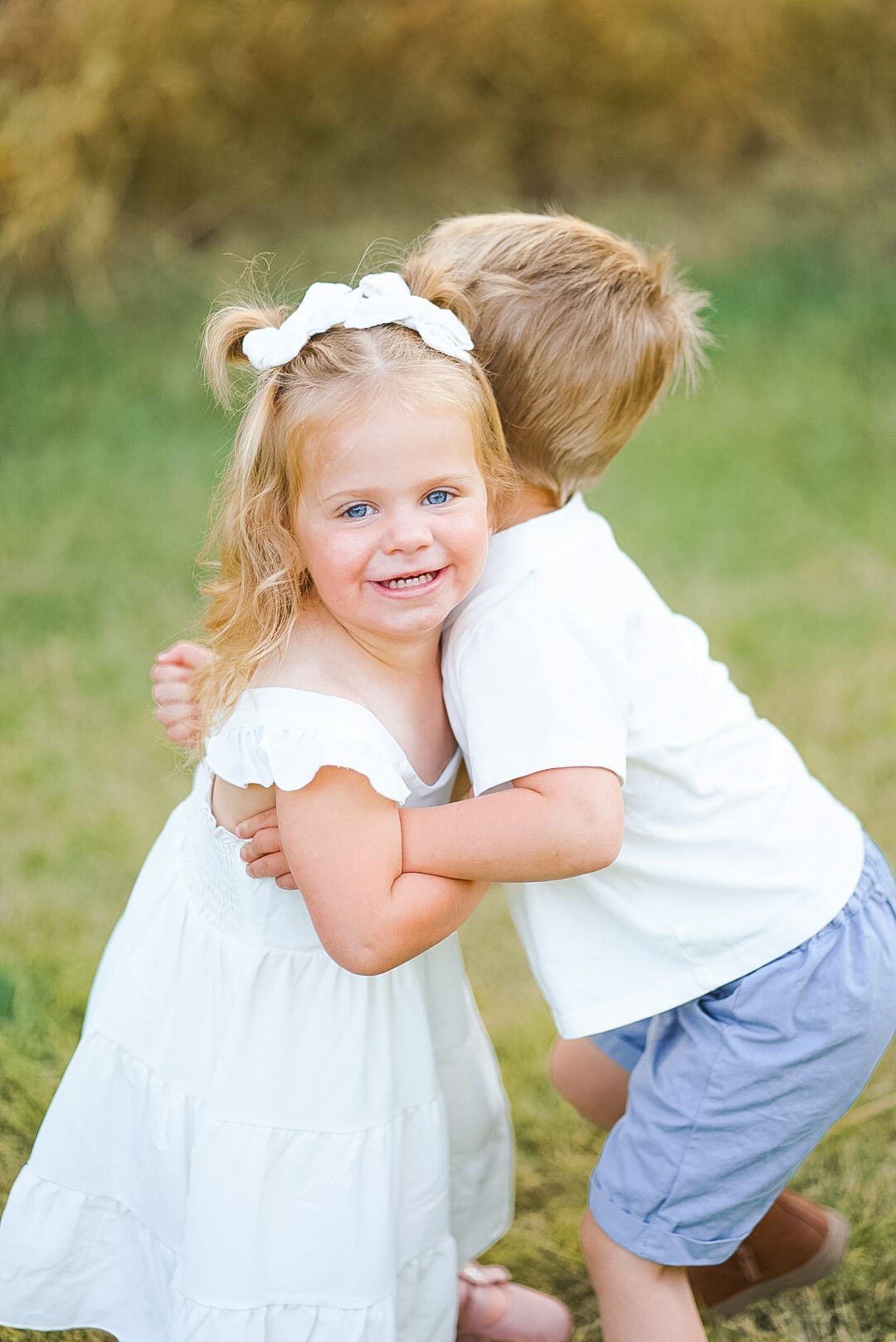 Two adorable blonde toddlers hugging each other in grassy outdoor setting wearing white outfits.