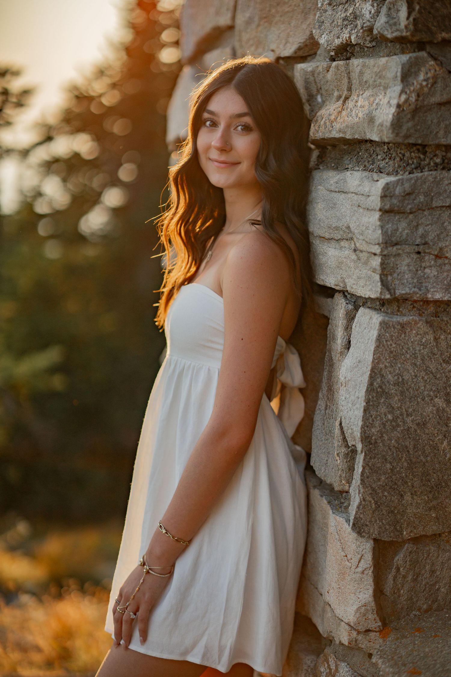 Teen girl in white sundress leaning against stone wall in warm golden sunset light.