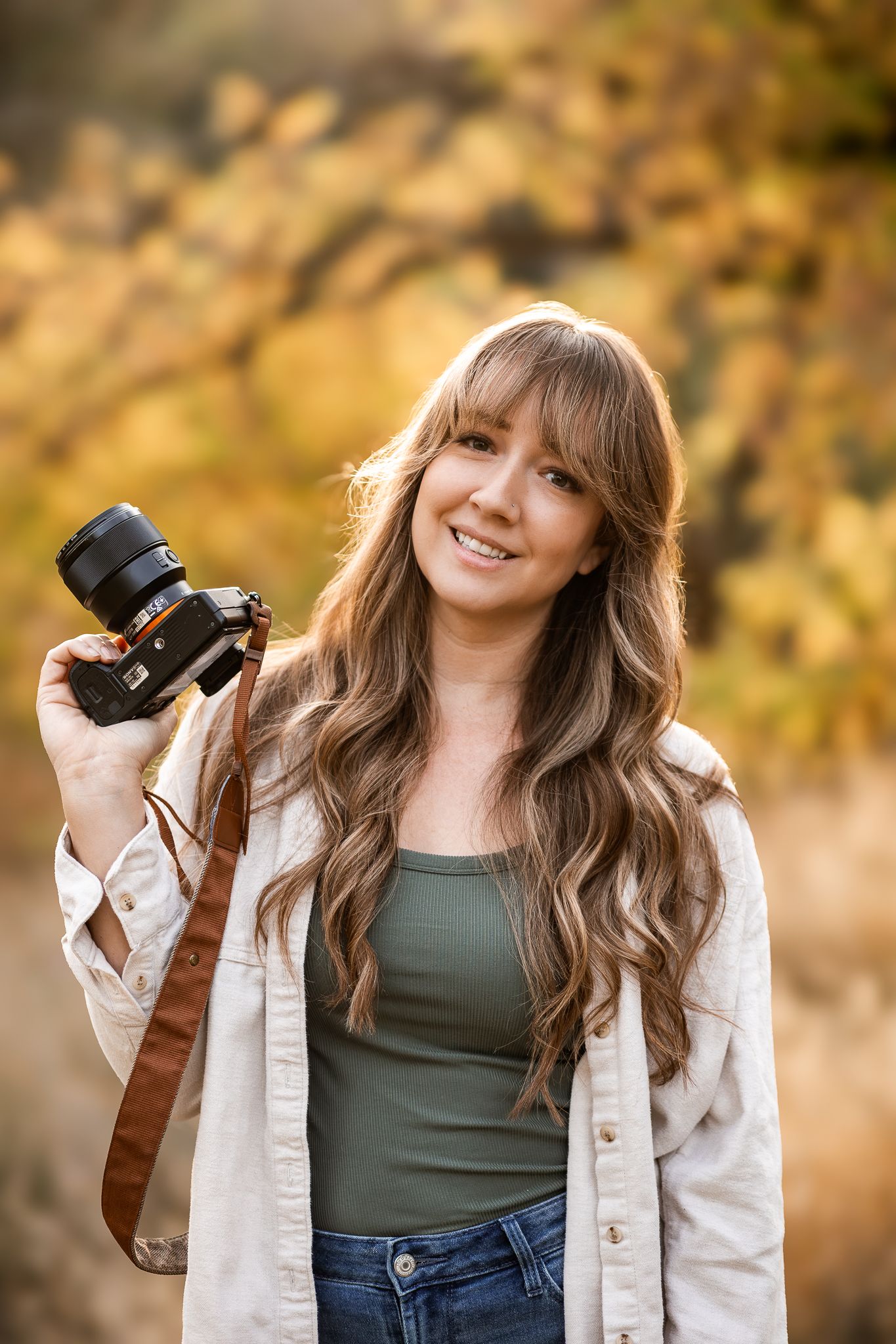 Smiling woman with long wavy hair holds a camera outdoors amid golden autumn foliage wearing jeans and a white shirt.
