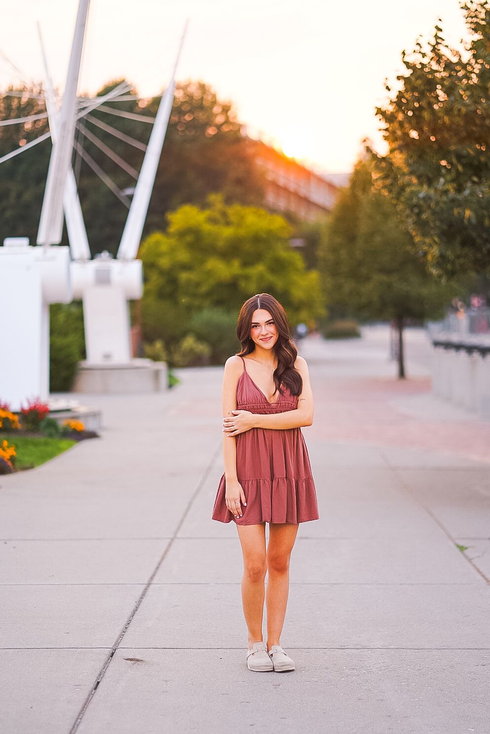 Young woman in burgundy sundress standing on urban walkway at golden hour sunset.