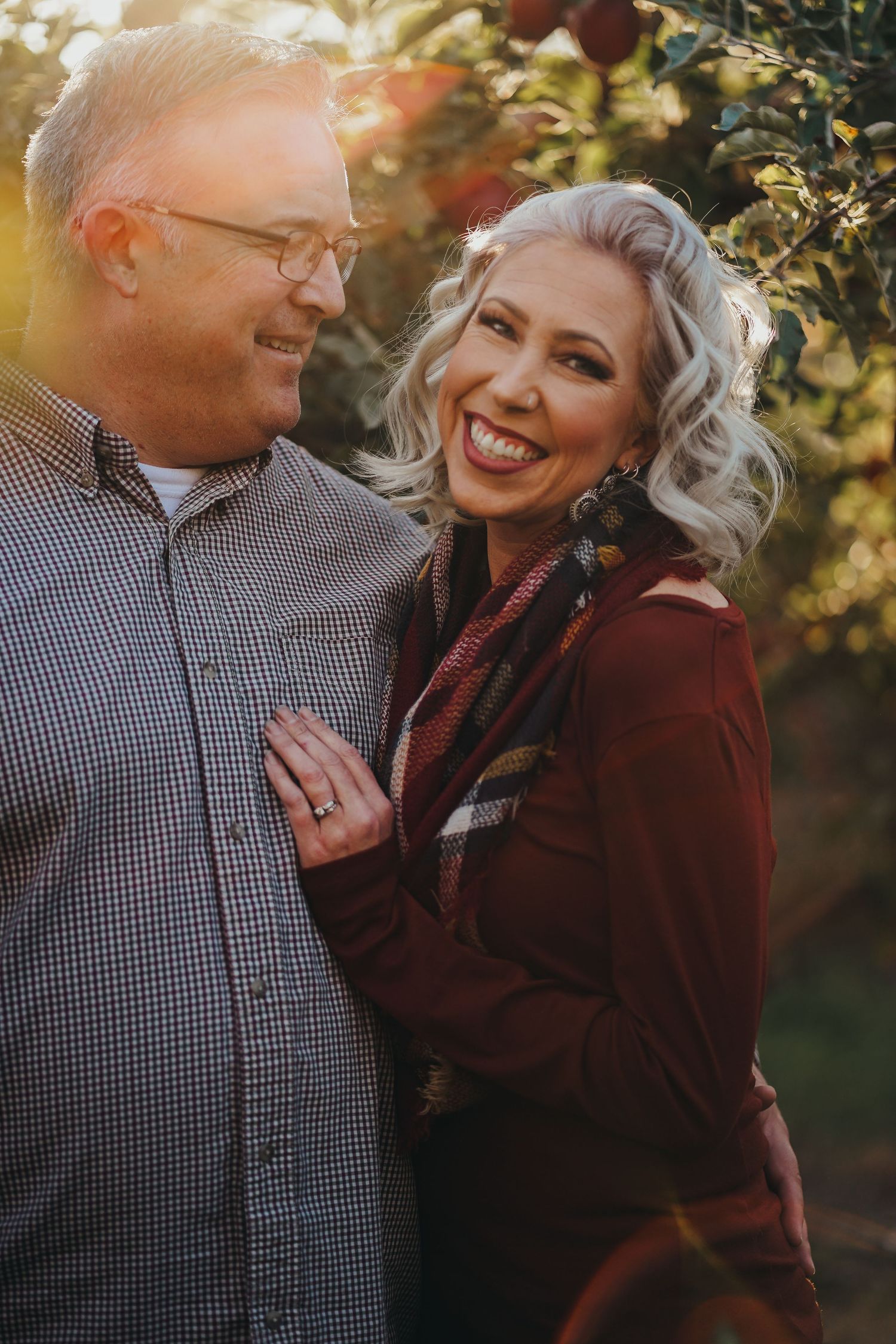 Happy couple posing in an apple orchard with golden sunlight, woman smiling brightly in burgundy.