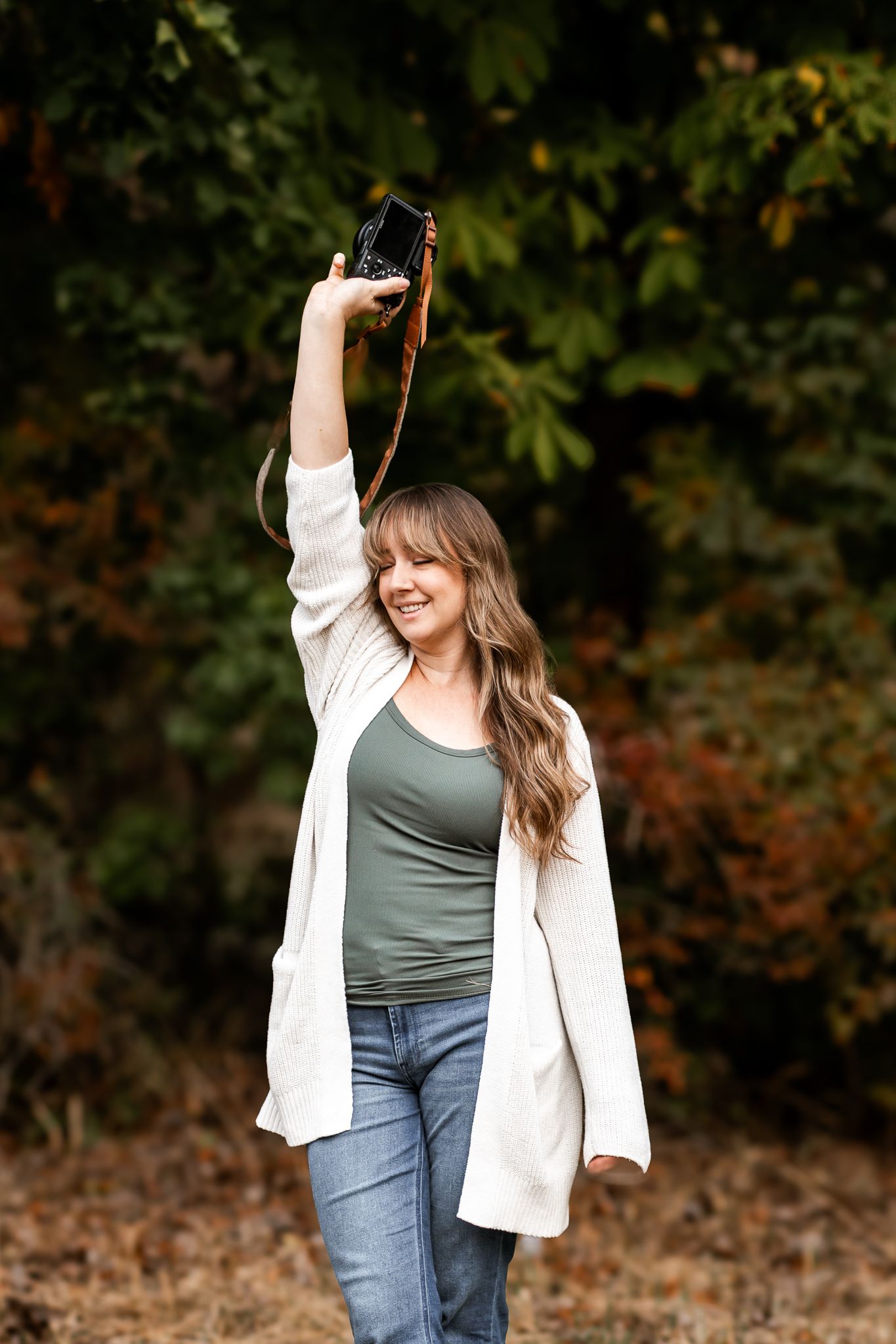 Woman in white cardigan holds vintage camera above head outdoors among autumn foliage.