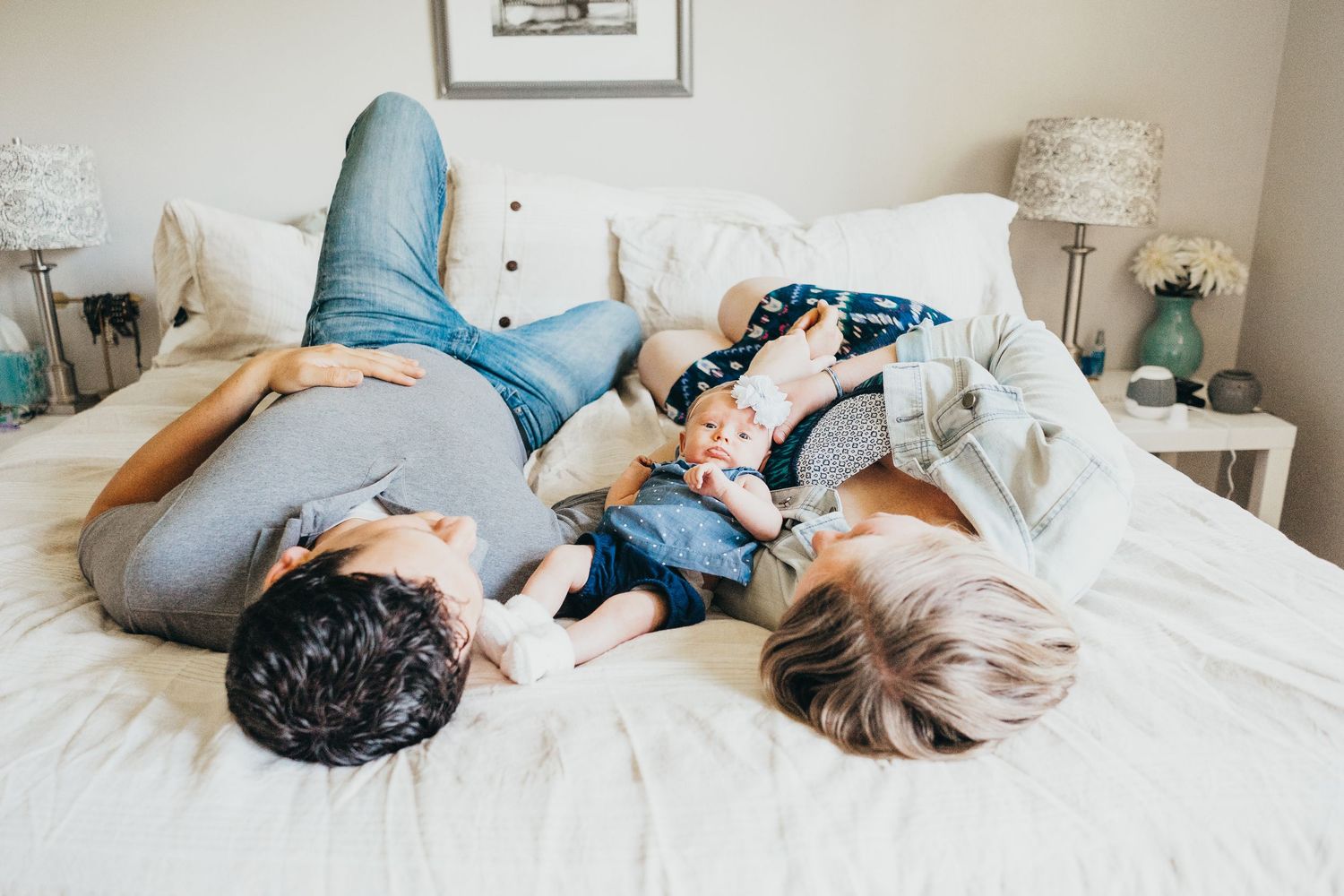 Parents lying on white bed with newborn baby between them in cozy home setting.