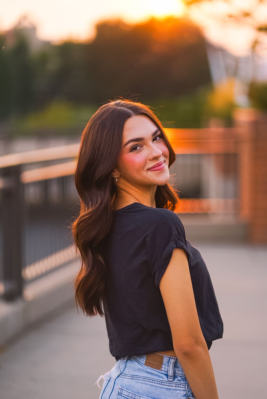 Smiling young woman in black top and jeans looking back over shoulder at golden sunset.