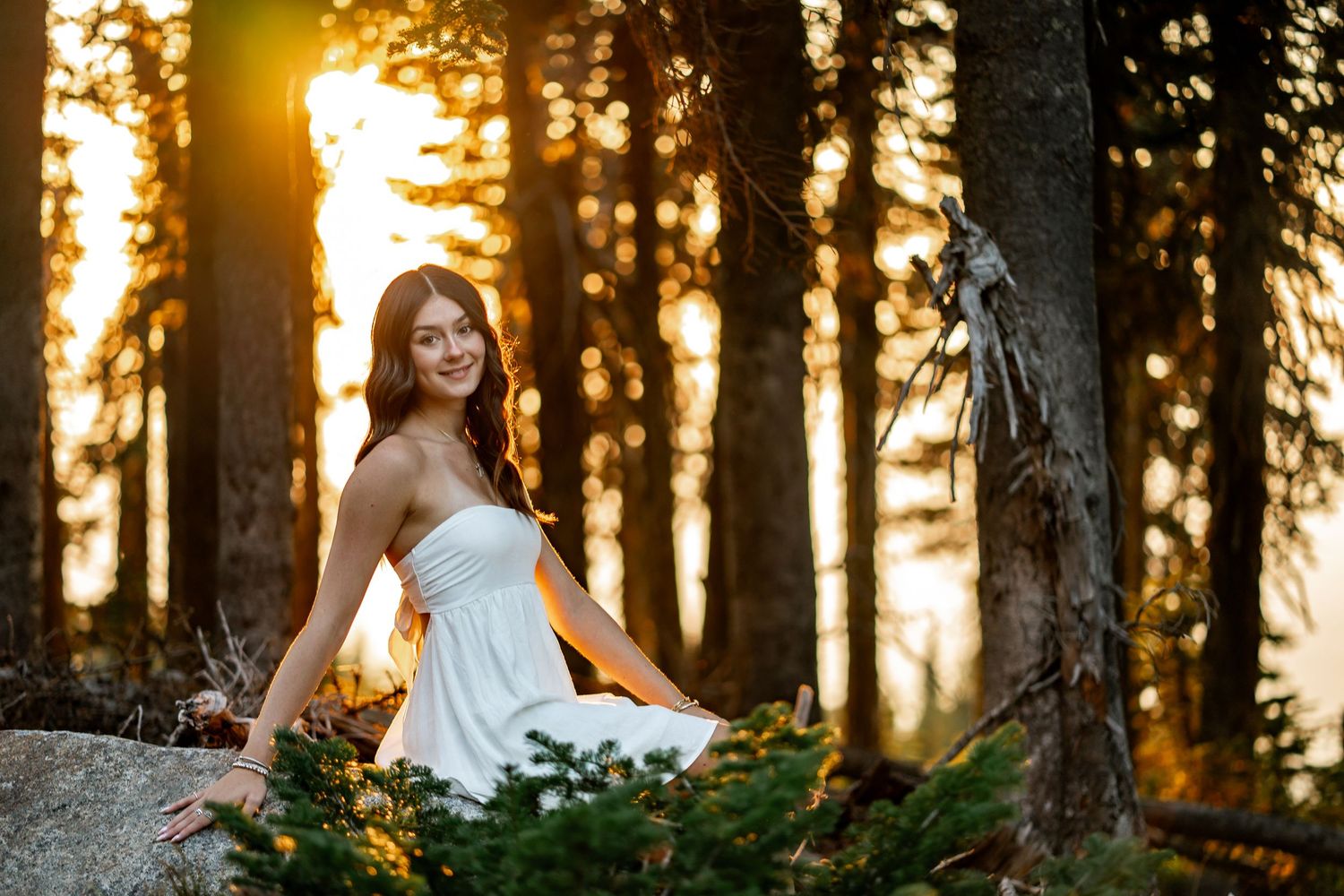 Smiling young woman in white strapless dress posing on rocks in glowing sunset forest.