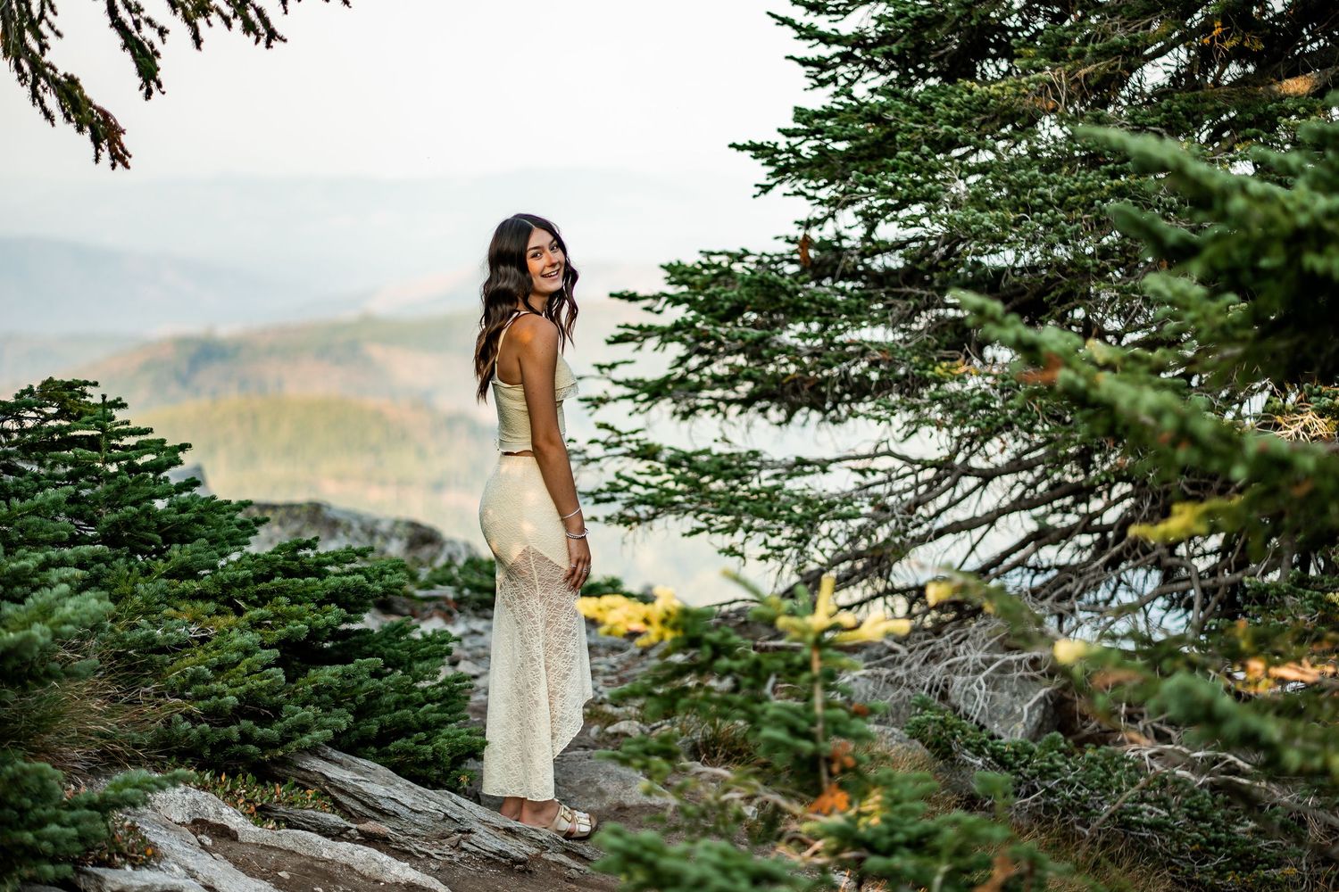 Elegant woman in white lace dress standing on rocky mountain overlook at dusk.