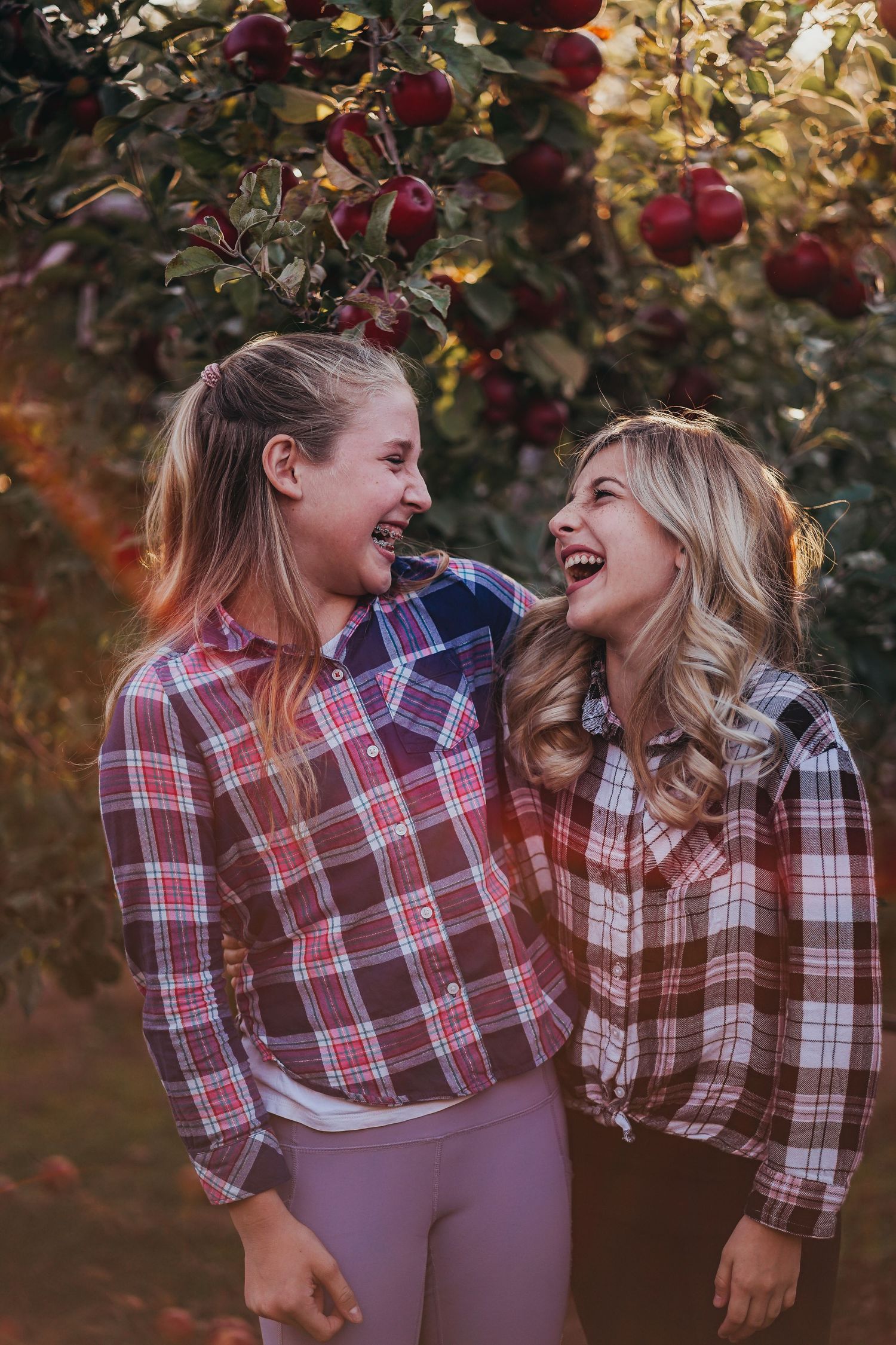 Two girls in matching plaid shirts laughing together under red apple tree branches.
