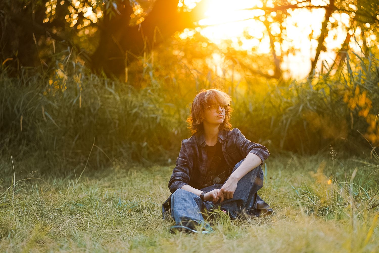 Young person with wavy hair sitting cross-legged in grass during golden sunset.