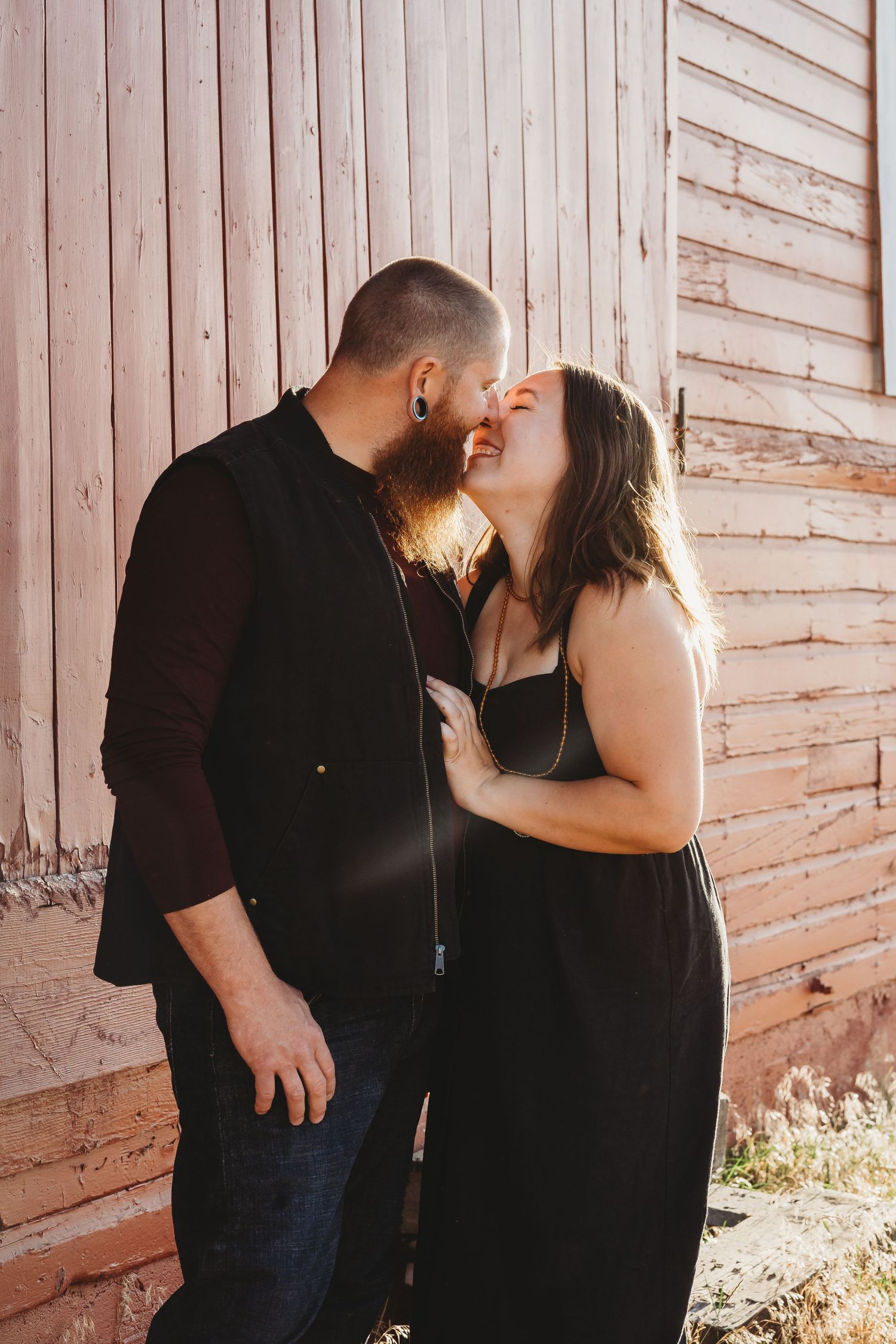 Couple sharing a romantic kiss outdoors against a rustic wooden barn wall in sunlight.