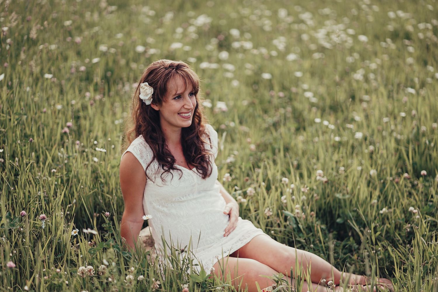 Pregnant woman in white dress sitting in a wildflower meadow with a flower in her hair.
