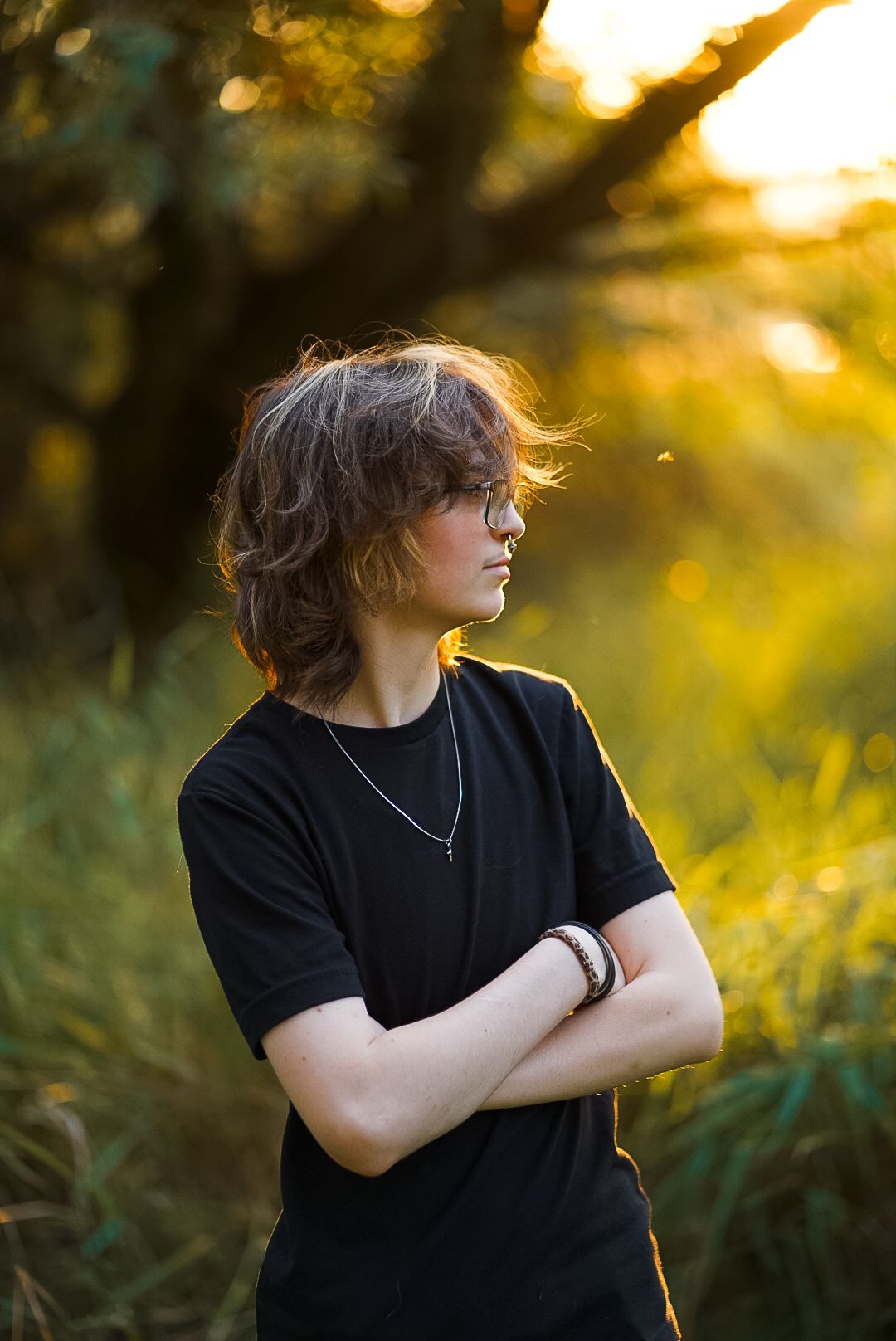 Teen with wavy hair and glasses standing arms crossed in golden sunset meadow.