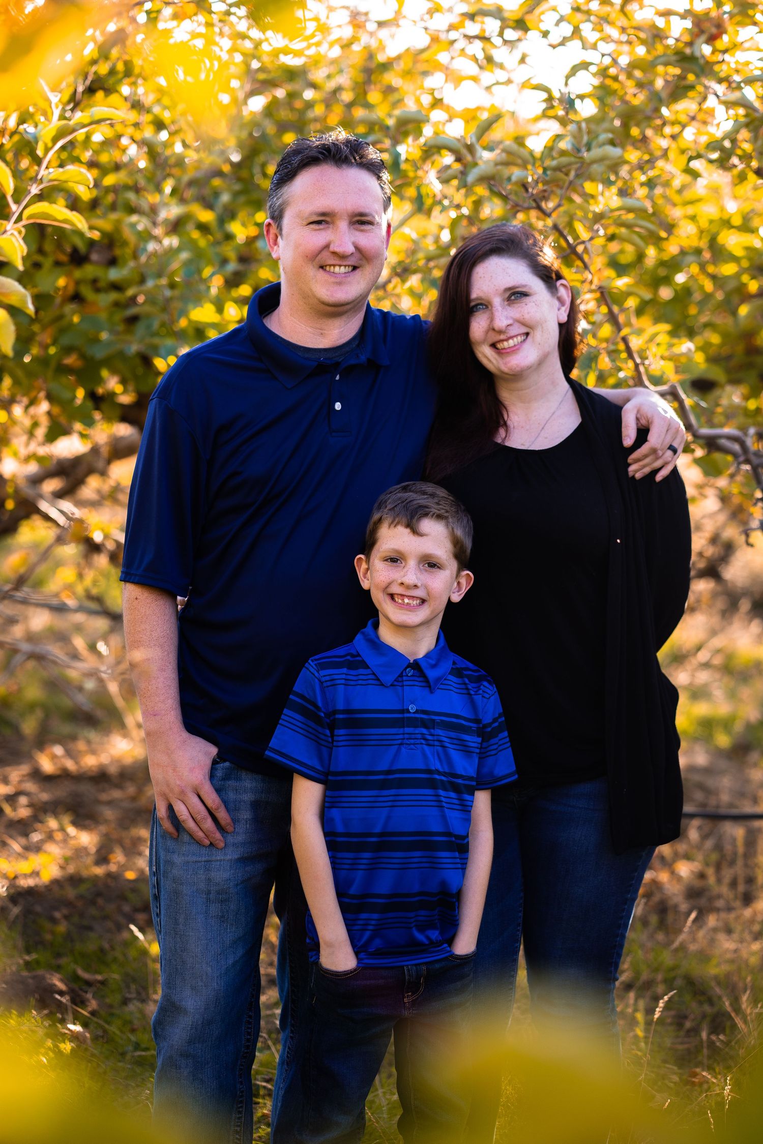 Smiling family of three posing together in golden sunlit apple orchard in autumn.