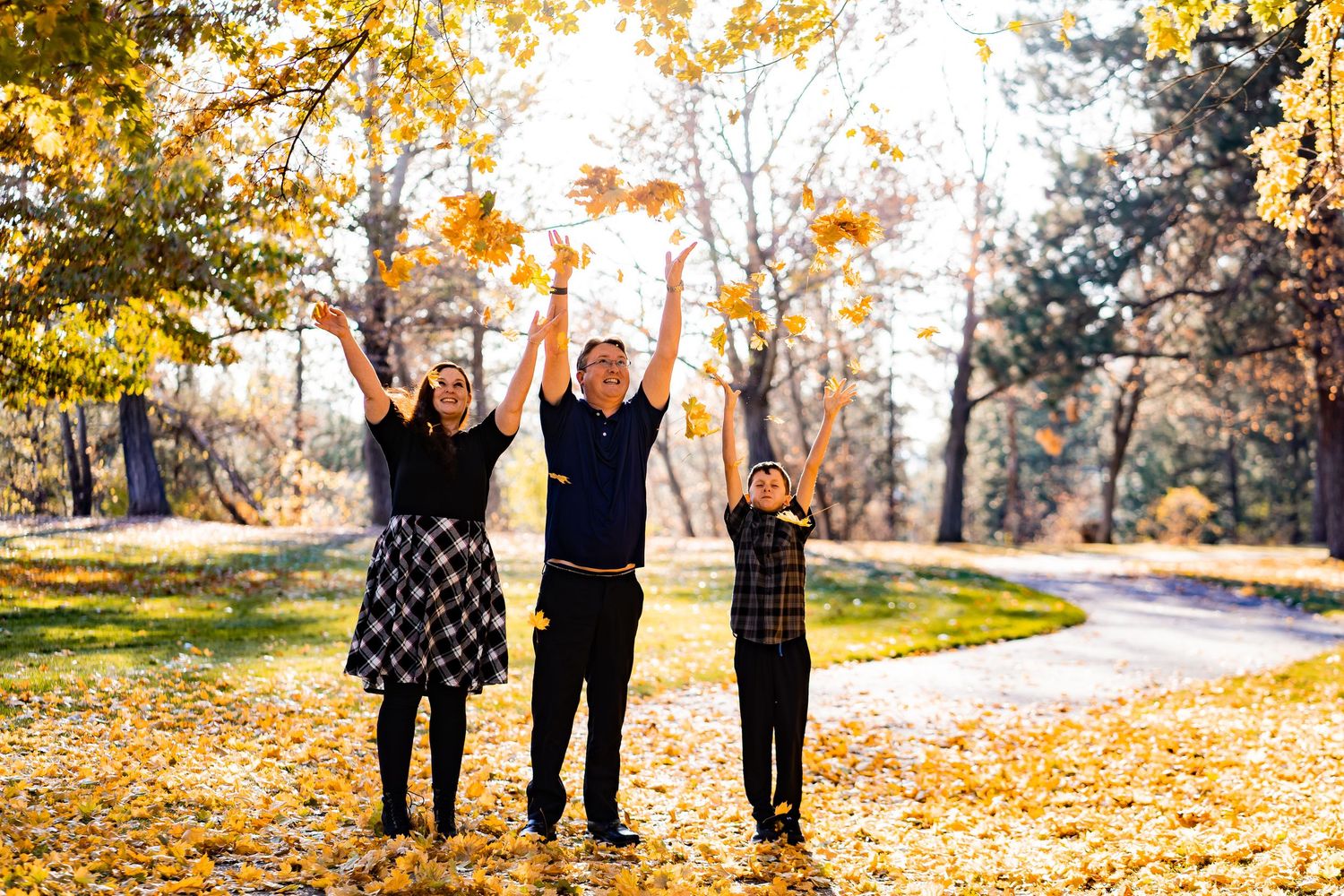 Family of three joyfully tossing autumn leaves in a park surrounded by golden fall foliage.