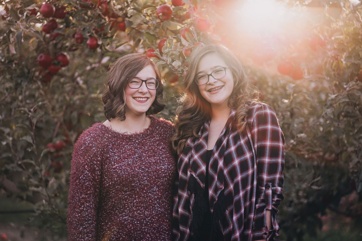 Two young women wearing glasses smiling together in front of apple trees in golden sunlight.
