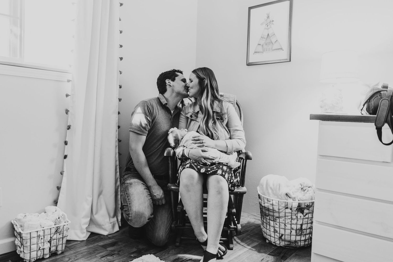 Black and white photo of couple kissing in nursery while holding newborn baby in rocking chair.