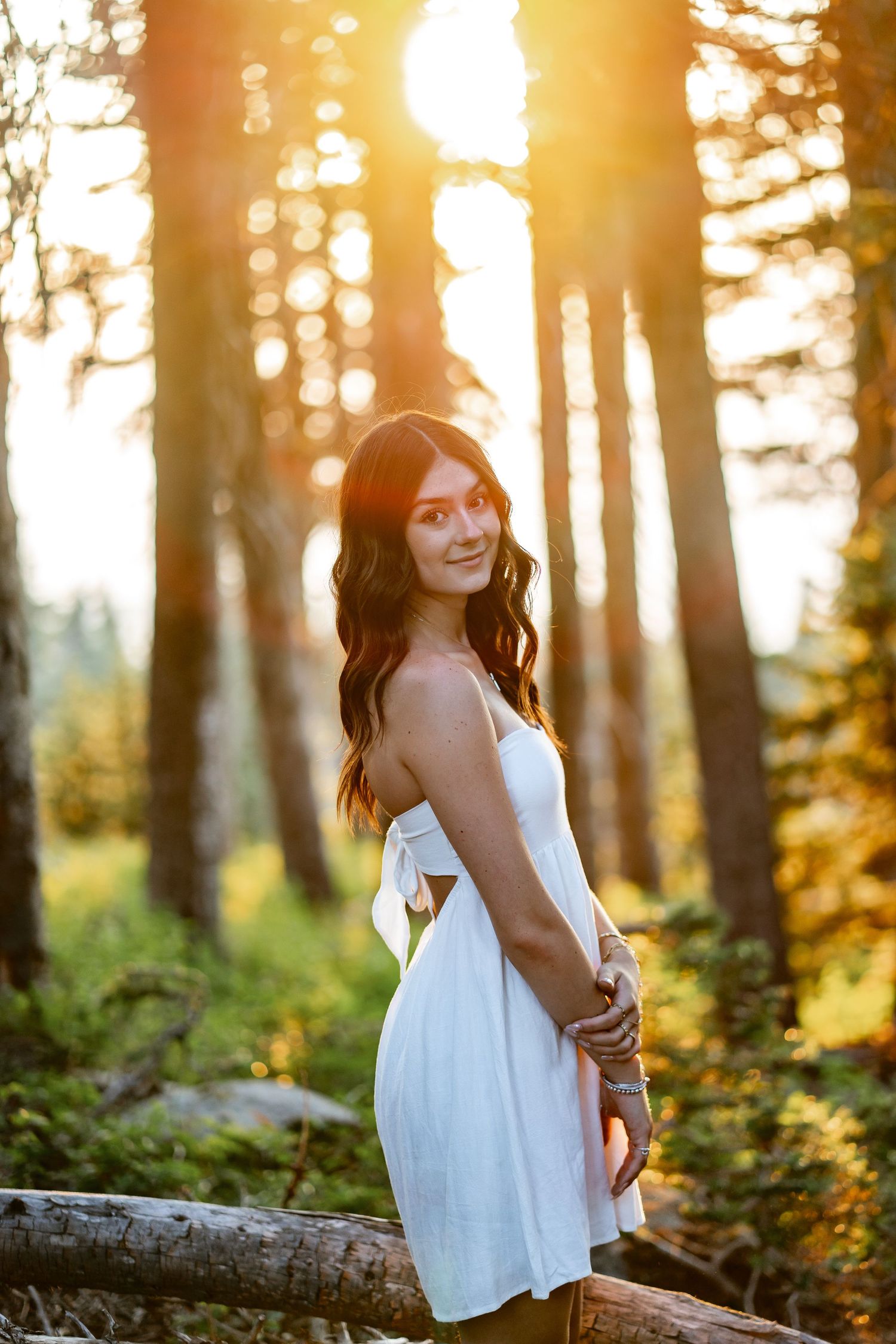 Young woman in white sundress standing by fallen log in radiant golden sunset forest.