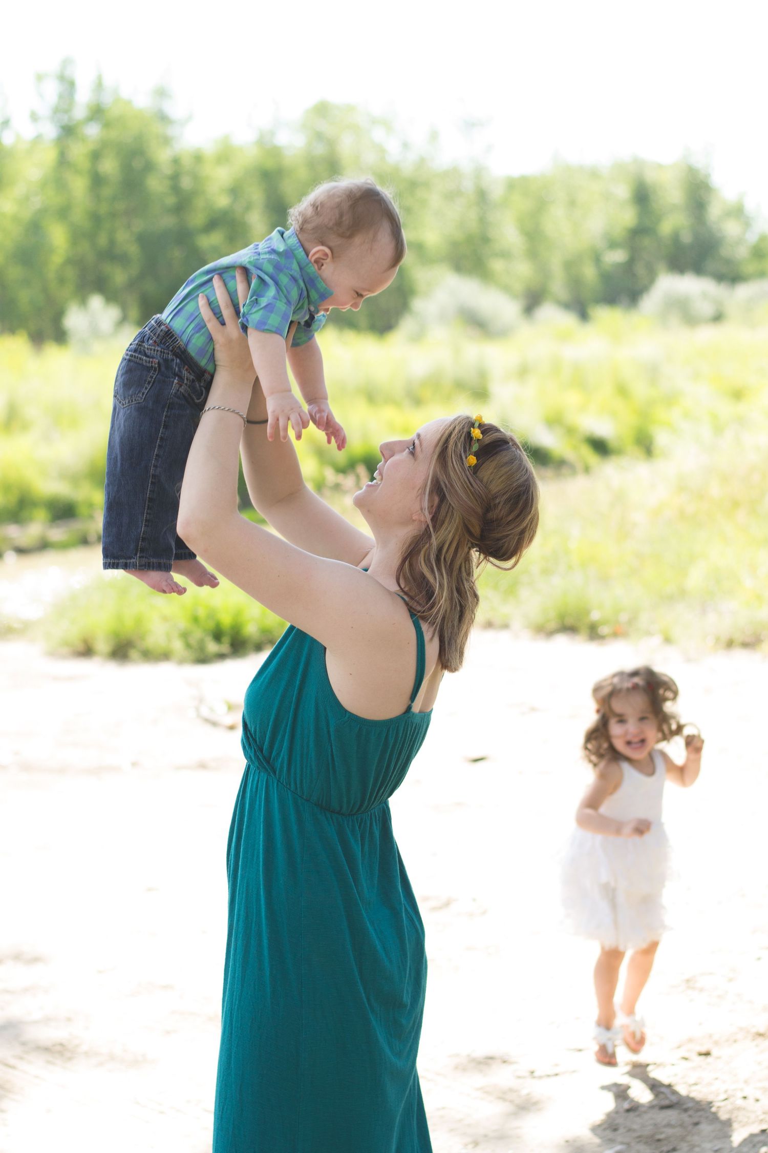 Mother in teal dress lifting smiling baby while toddler runs in background.