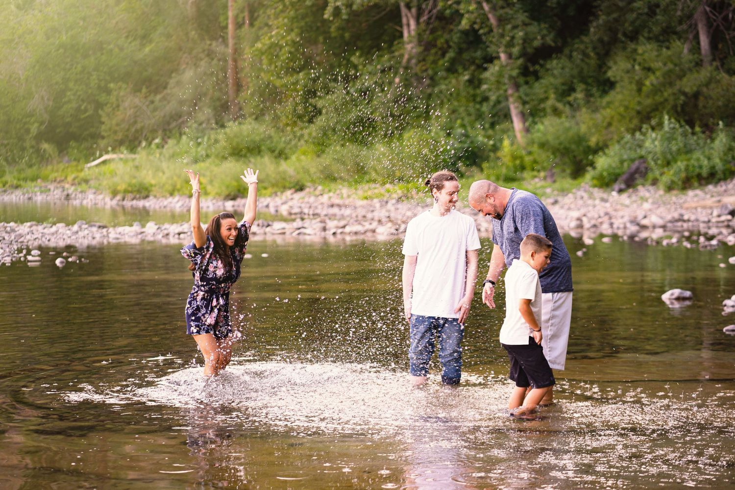 Family of four playing and splashing in a shallow river surrounded by lush greenery.