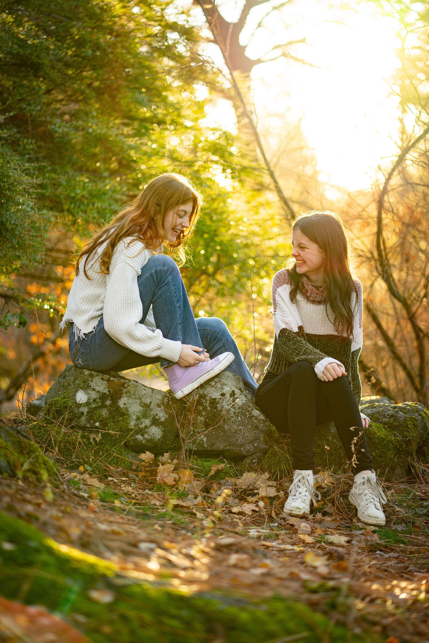 Two teenage girls sitting on rocks in a sunlit autumn forest, smiling and talking together.